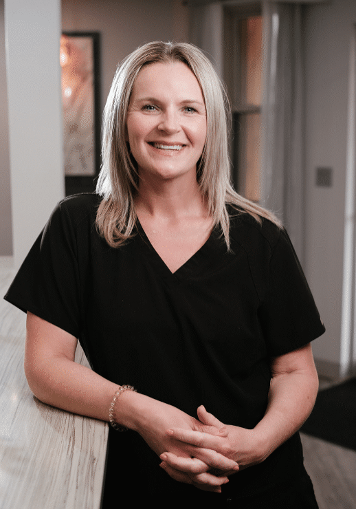 Smiling woman in black scrubs standing at a light wood counter in an indoor office setting, hands folded on the counter.
