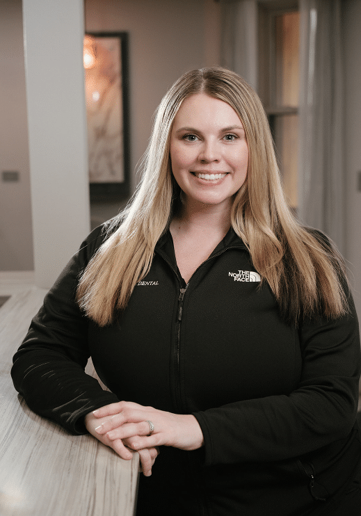 Smiling woman with long blonde hair in a black jacket leaning on a marble counter in a dental office.